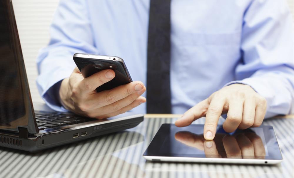 Ingezoomde foto van iemand die aan een bureau zit en tegelijkertijd op zijn laptop, mobile telefoon en tablet zit.