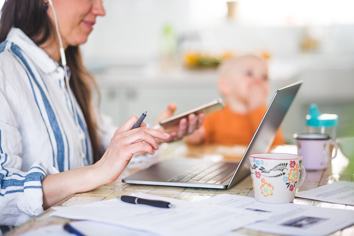 Foto van een persoon die aan het werk is aan de keukentafel. De persoon heeft oortjes in en een telefoon in de hand en op tafel staat een laptop en liggen er papieren. Op de achtergrond zit een kindje in een kinderstoel te eten.