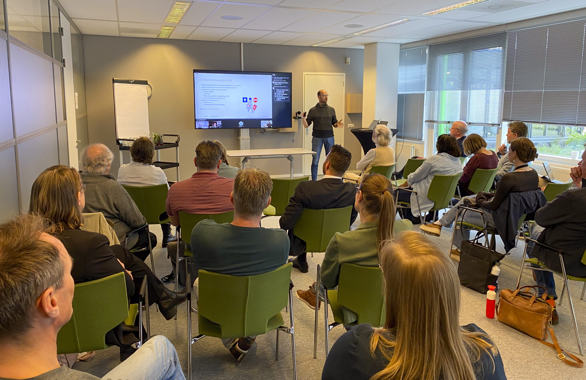 Een groep personen zitten op groene stoelen te kijken naar een presentatie tijdens de twaalfde werksessie van de zib-transitie